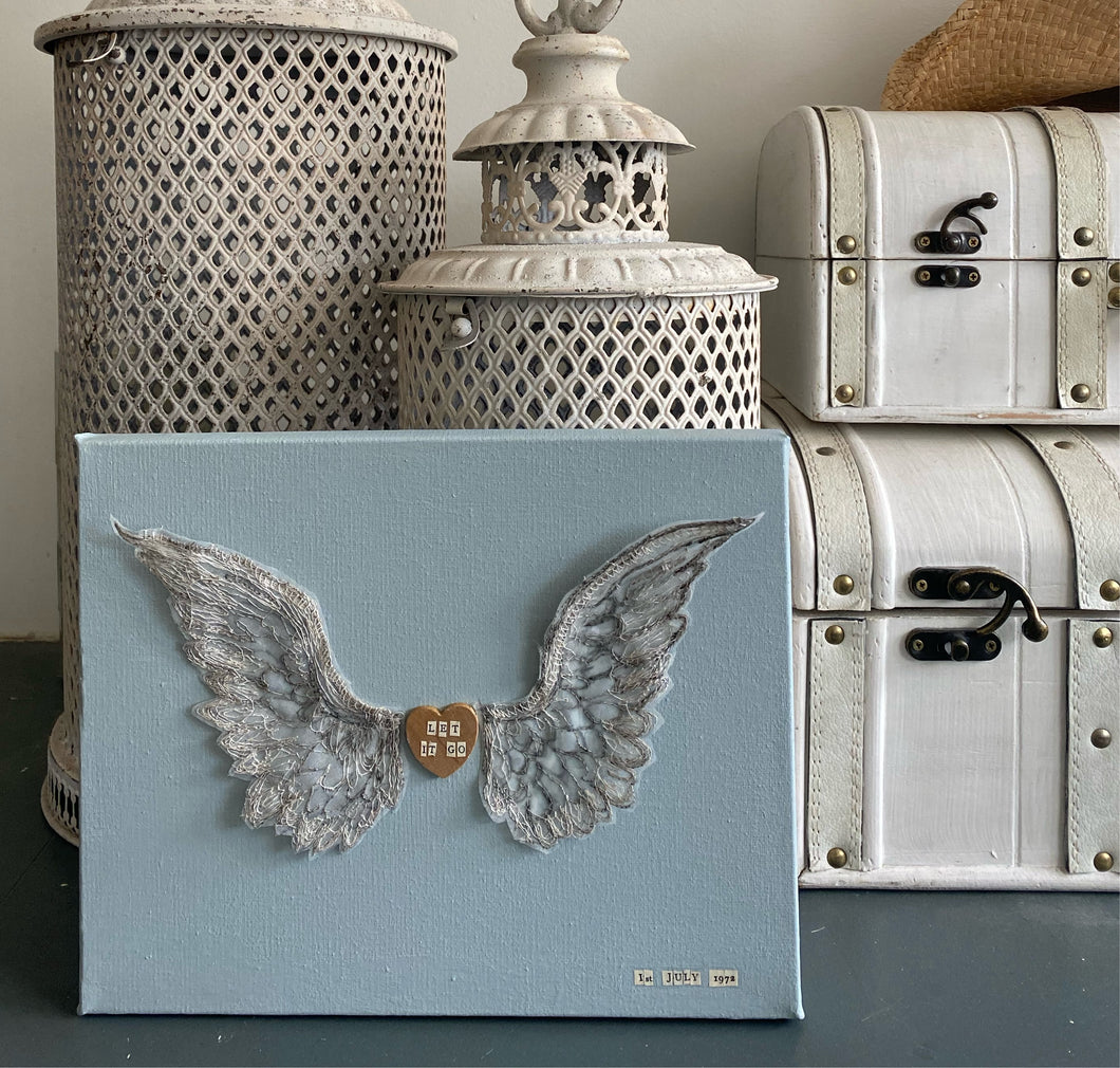 Personalised embroidered wings on a duck egg blue canvas with a wooden heart featuring names, dates, or quotes.